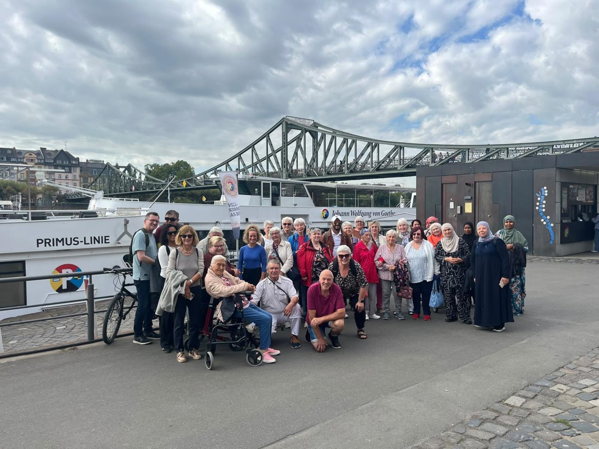 Eine Gruppe von 32 Seniorinnen und Senioren mit 3 begleitenden Personen posiert vor dem Schiff "Johann Wolfgang von Goethe" und dem Eisernen Steg am Main.