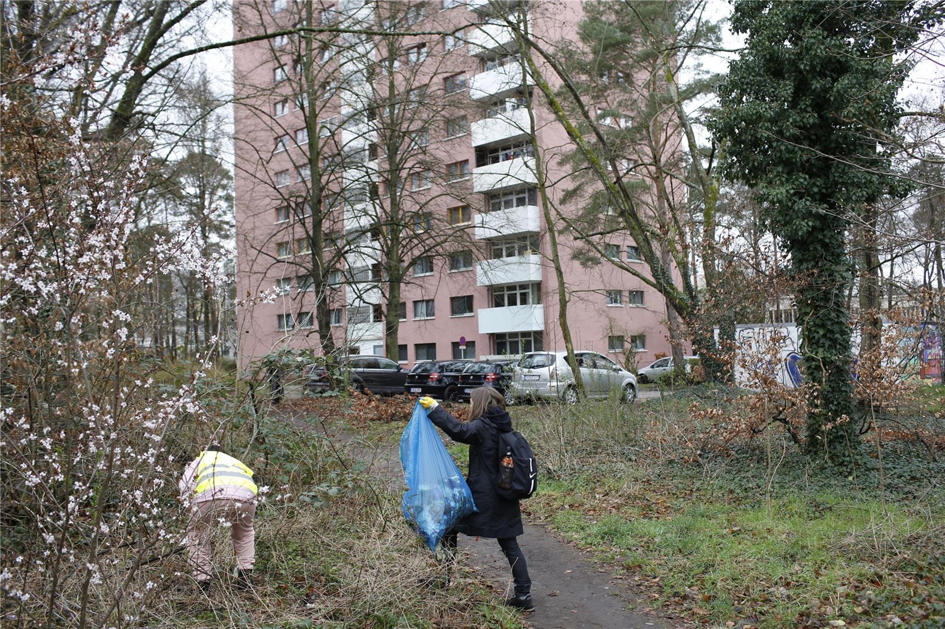 Eine Frau mit Handschuhen trägt einen großen Müllsack, eine andere Person in Warnweste bückt sich nach Müll im Grünstreifen vor einem großen Hochhaus